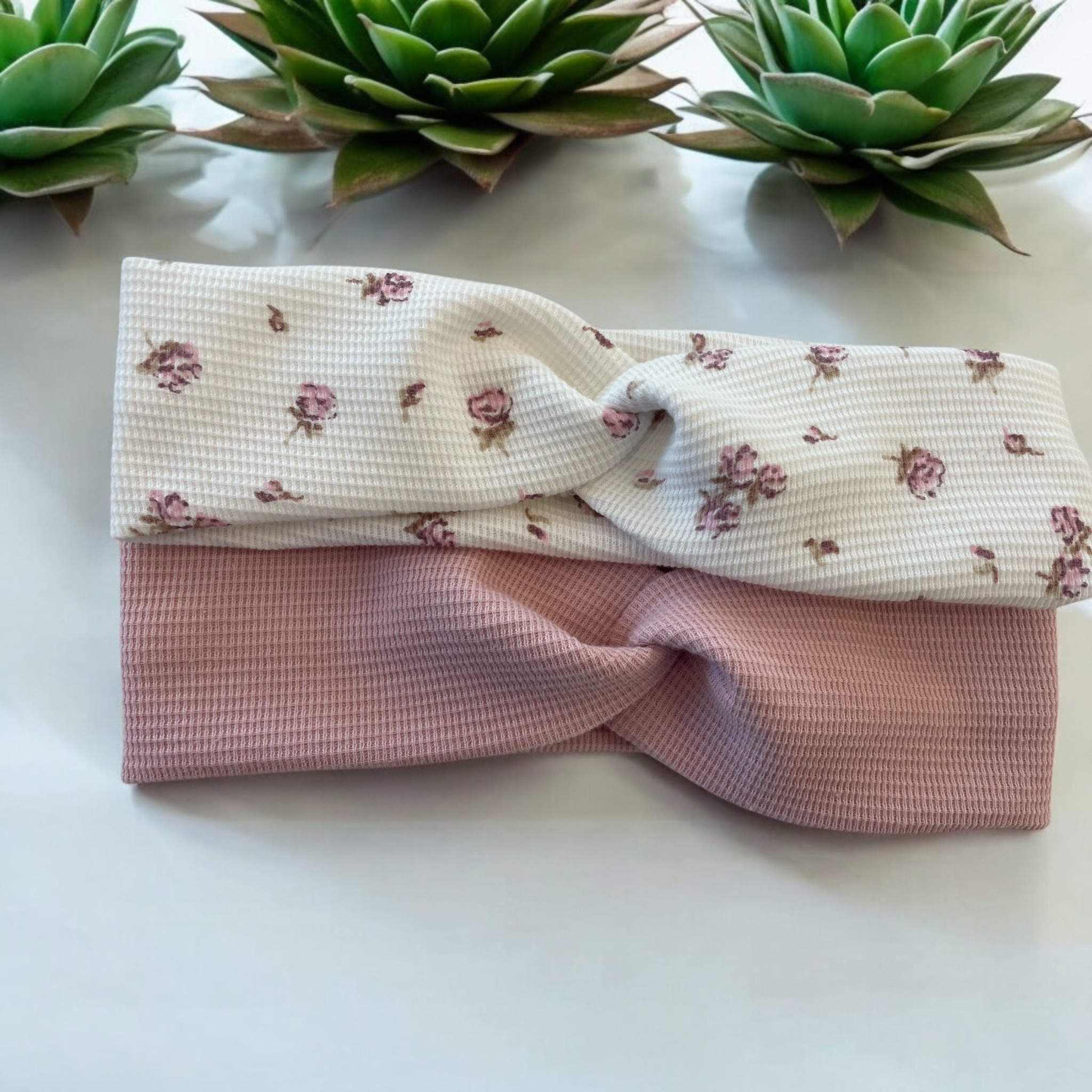 Dwie opaski do włosów z materiału wafelkowego: różowa i biała w kwiaty, leżące na białym tle, z sukulentami w tle. Ujęcie z góry, miękkie światło.