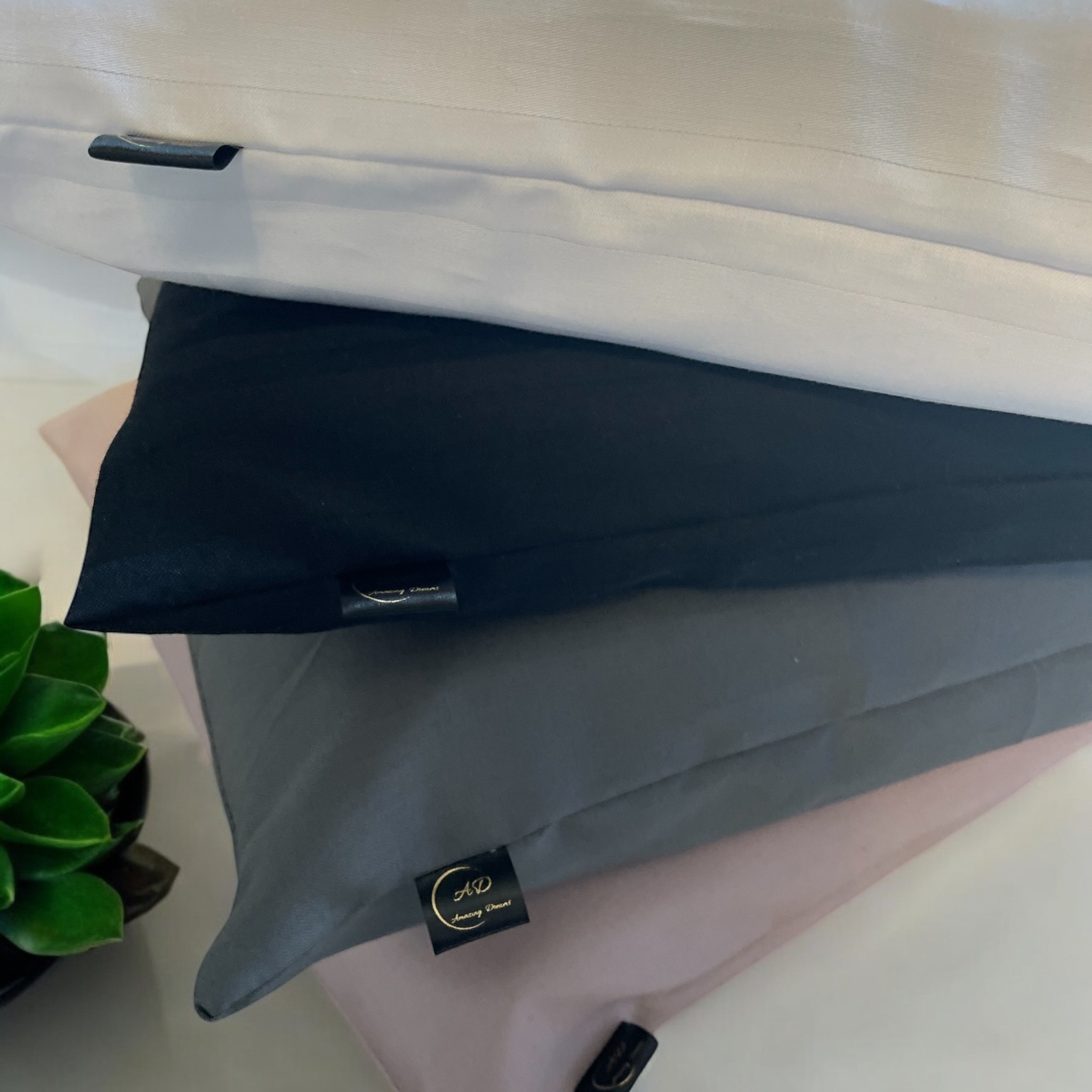 Stos poduszek dekoracyjnych w odcieniach bieli, czerni, szarości i pudrowego różu, z dyskretnymi metkami marki, obok doniczki z zieloną rośliną.