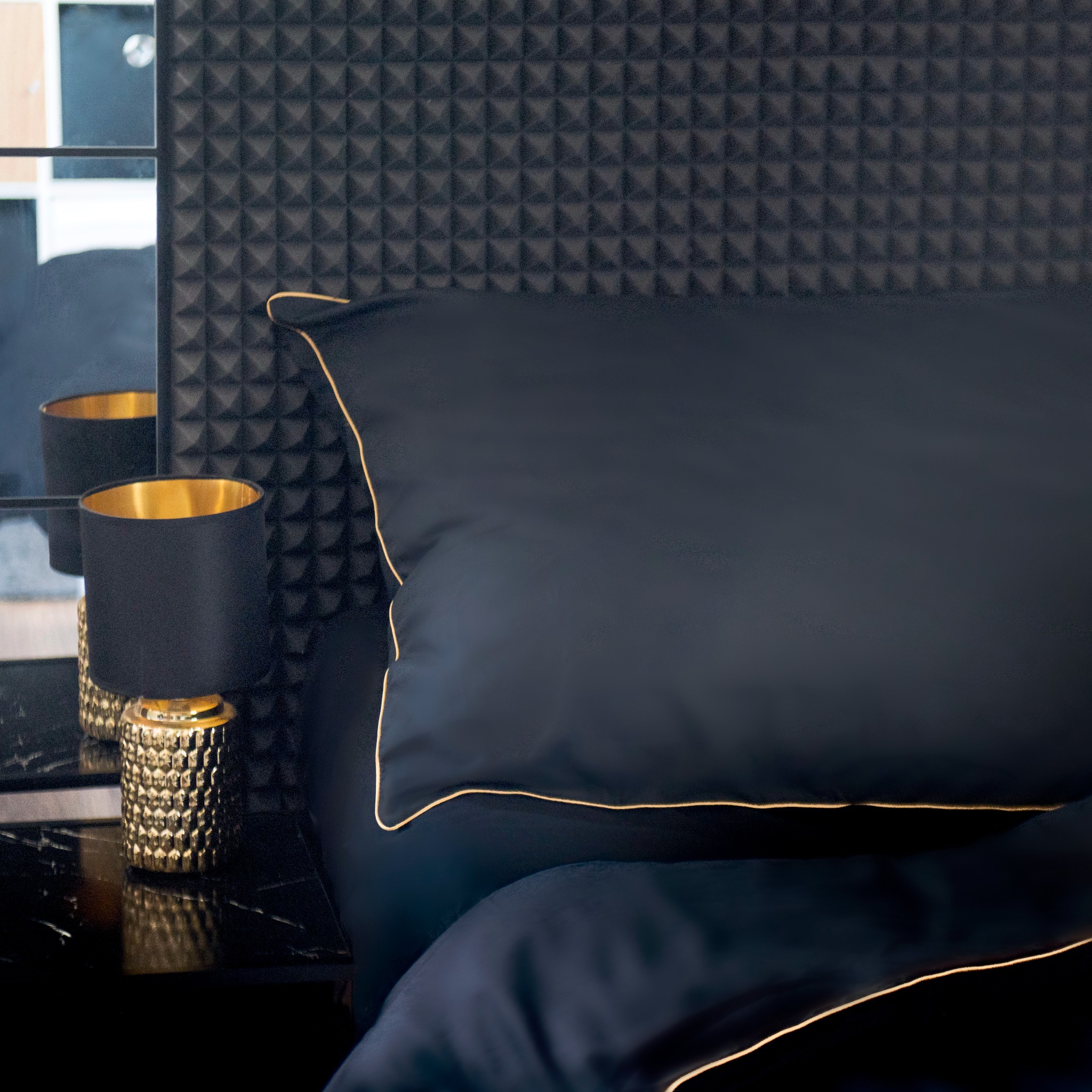 Elegancka, ciemna pościel z złotą lamówką na poduszce i kołdrze, obok lampka nocna z lustrzanym odbiciem, na tle ściany z geometrycznym wzorem.