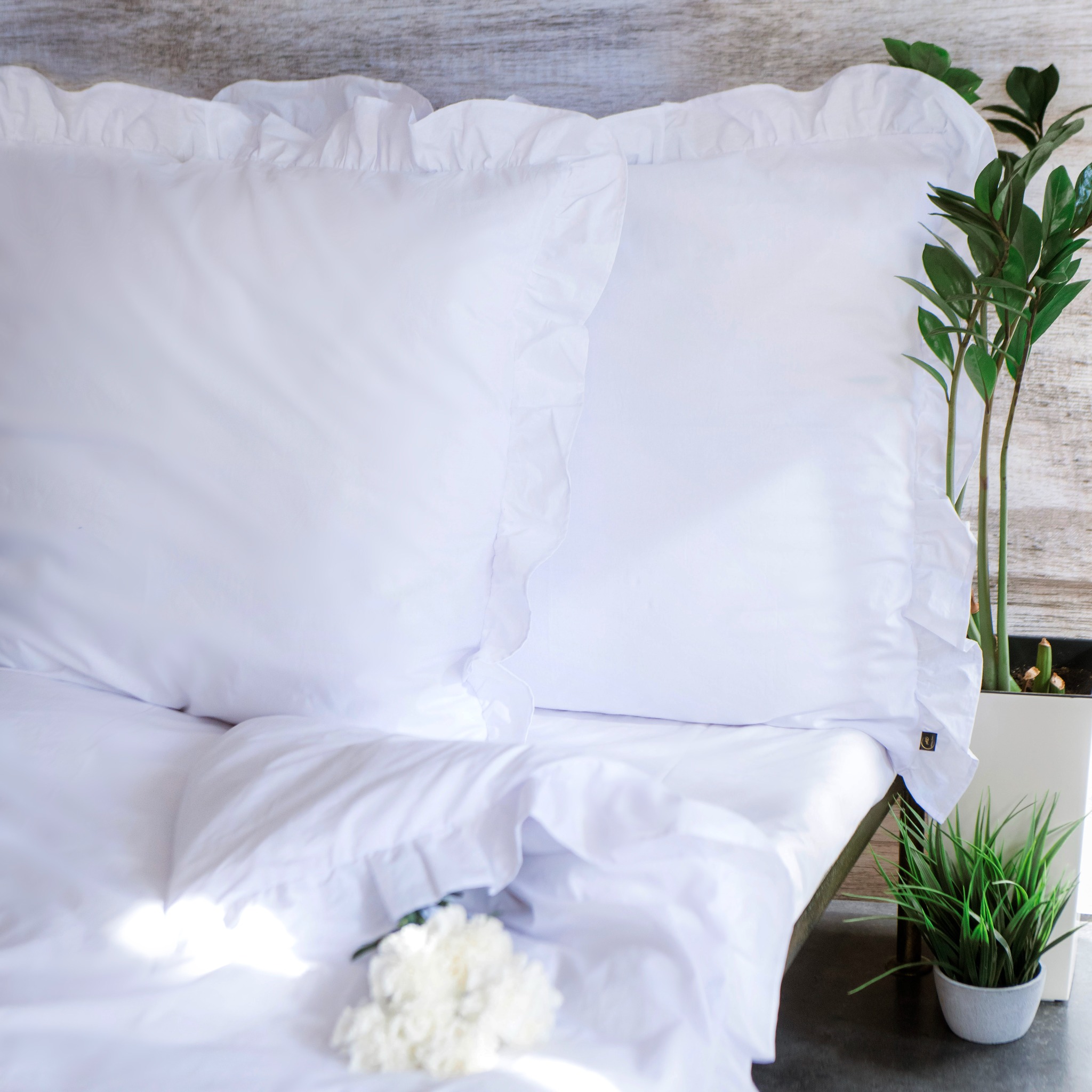 Biała pościel z falbankami na poduszkach, uzupełniona kwiatami i rośliną doniczkową, tworzy minimalistyczną i elegancką aranżację sypialni.
