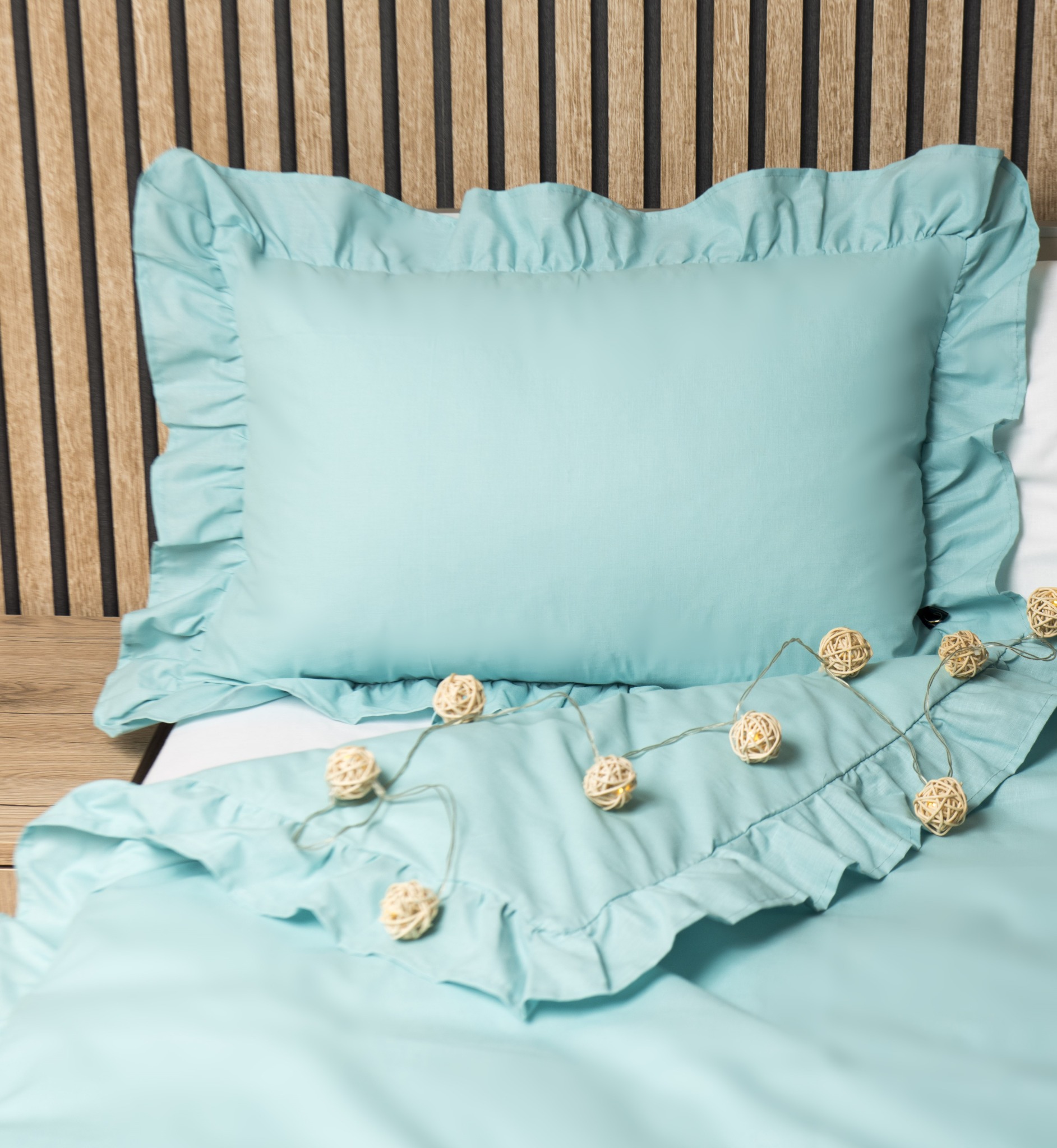 Turkusowa pościel z falbankami na łóżku z drewnianym zagłówkiem, oświetlona girlandą z rattanowych kul. Estetyczne ujęcie gotowego produktu.
