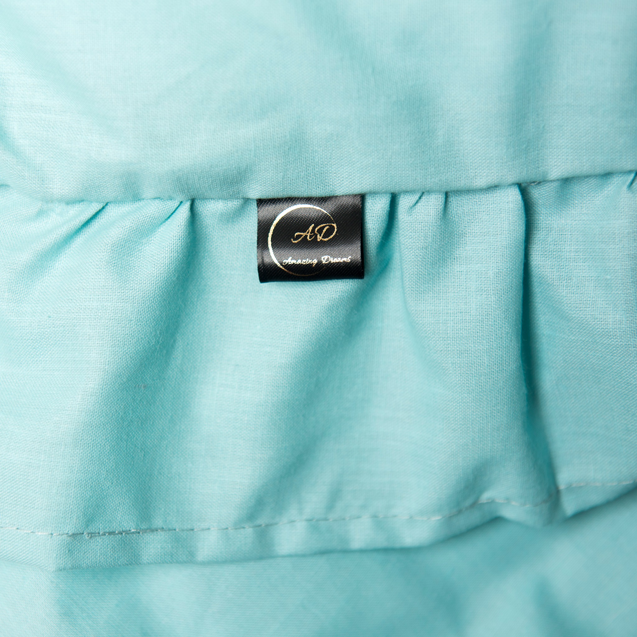 Turkusowa tkanina z falbanką i czarną metką z logo 'AD Amazing Dresses' w złotym kolorze. Detal wykończenia odzieży dziecięcej.