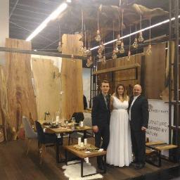 Handicraft - Ekspozycja na targach mebli: stół z blatem drewnianym i krzesłami, tło z dużych desek drewnianych, oświetlenie z żarówek Edisona, oraz trzy osoby w eleganckich strojach pozujące do zdjęcia.