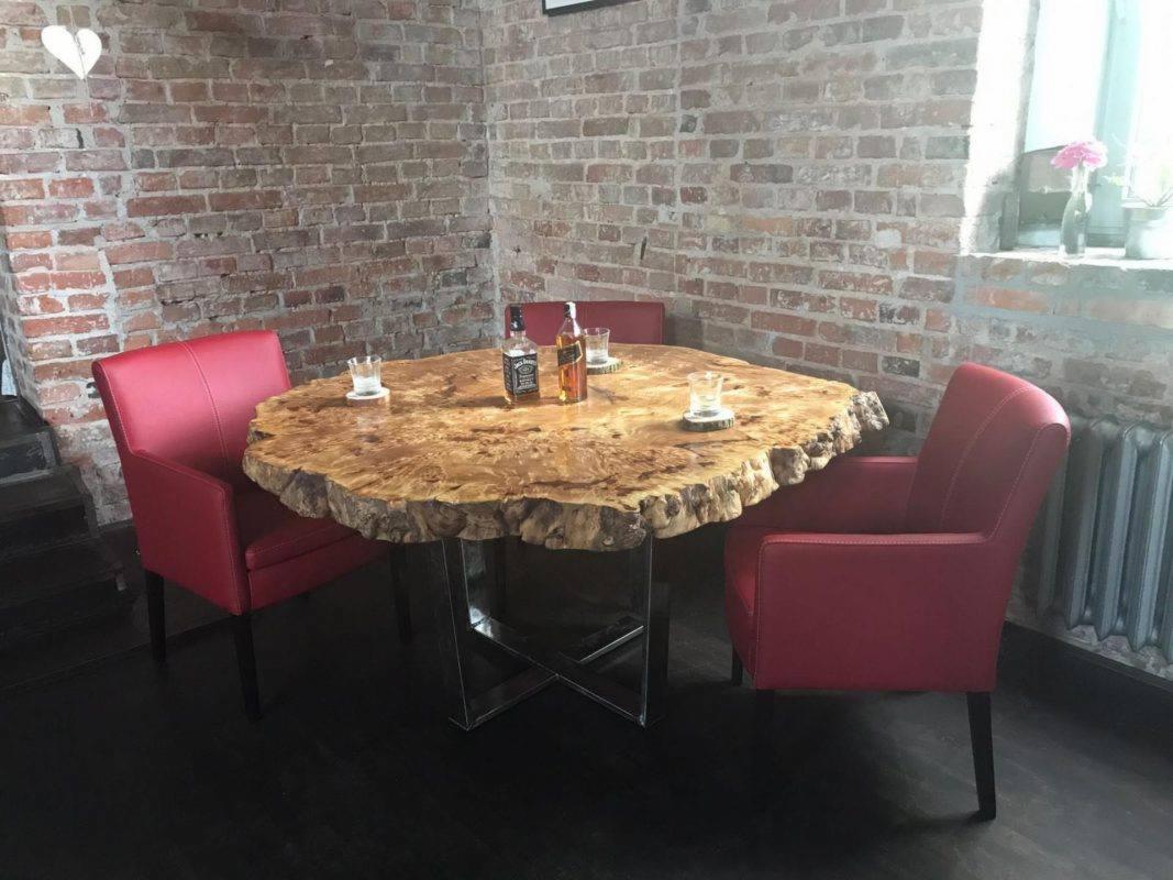 Stół z blatem z naturalnego drewna o nieregularnych krawędziach, wsparty na metalowej konstrukcji, otoczony trzema czerwonymi krzesłami, na blacie szklanki i butelki alkoholu, w tle ceglana ściana.