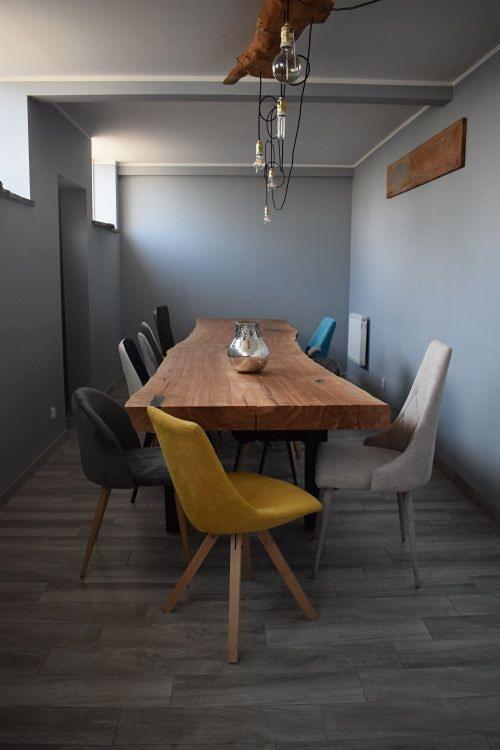 Długi stół z litego drewna z nieregularnymi krawędziami, otoczony różnymi krzesłami w odcieniach szarości, żółci i turkusu, oświetlony lampą wiszącą z surowego drewna i żarówek na kablach.