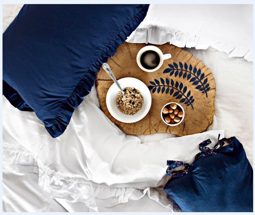 Śniadanie w łóżku: owsianka z łyżeczką, kawa i orzechy na drewnianej tacy z motywem liści na białej pościeli z falbankami i granatowych poduszkach.
