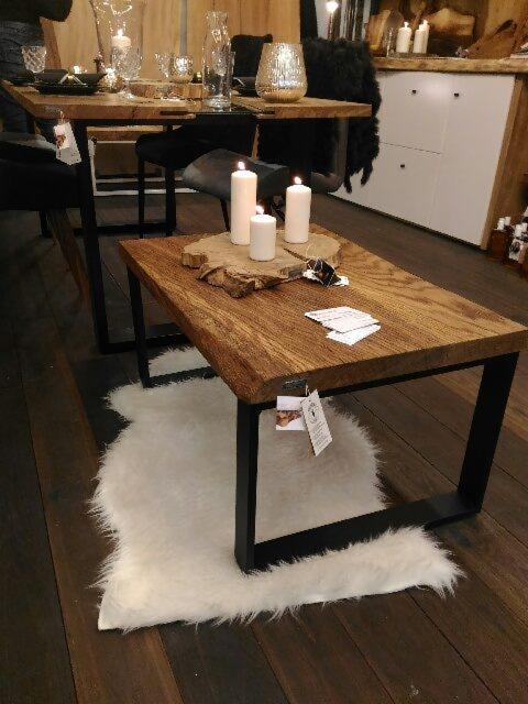 Stolik kawowy z blatem z litego drewna o nieregularnych krawędziach, na metalowych nogach w kształcie litery U, ustawiony na białej, puszystej skórze owczej, z zapalonymi świecami i drewnianą...