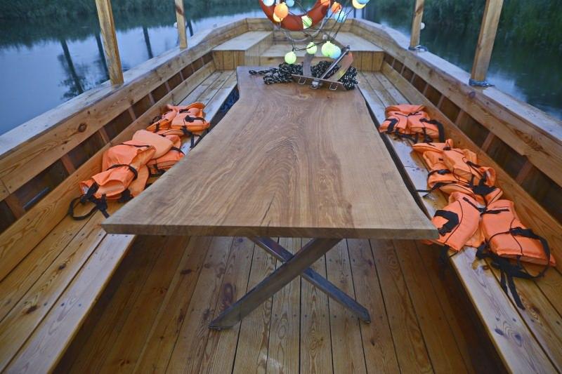 Drewniany blat stołu z nieregularnymi krawędziami, osadzony na metalowych nogach w kształcie litery X, umieszczony wewnątrz drewnianej łodzi z pomarańczowymi kamizelkami ratunkowymi po bokach...