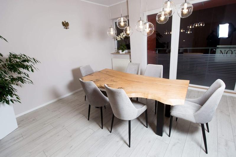 Stół z blatem z litego drewna o nieregularnym brzegu, wsparty na czarnych, prostych nogach, otoczony sześcioma szarymi krzesłami tapicerowanymi w nowoczesnym wnętrzu z jasną podłogą i wiszącymi...