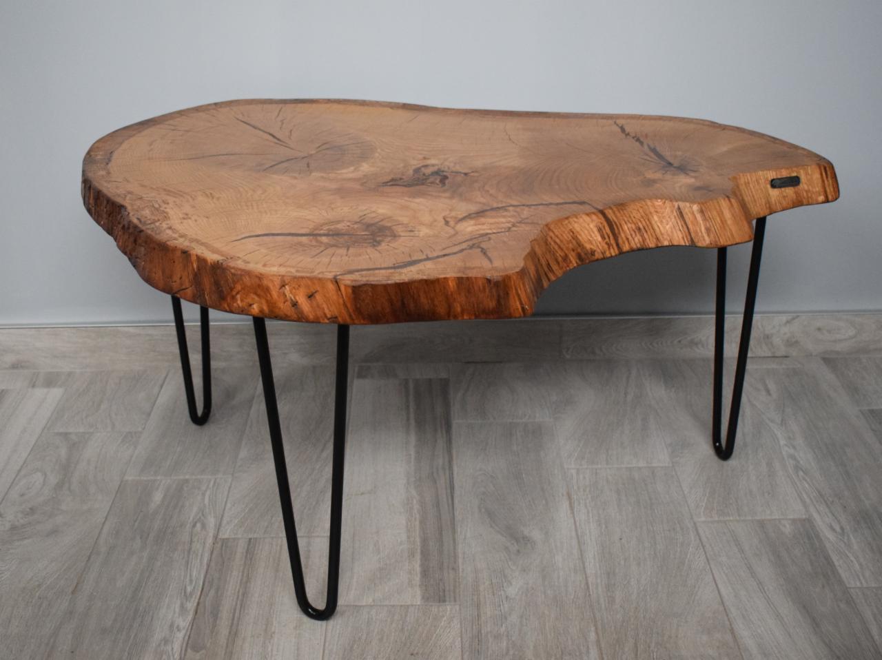 Stolik kawowy z blatem z litego drewna o nieregularnym kształcie, z widocznymi słojami i naturalną krawędzią, wsparty na trzech metalowych, czarnych nogach typu hairpin.