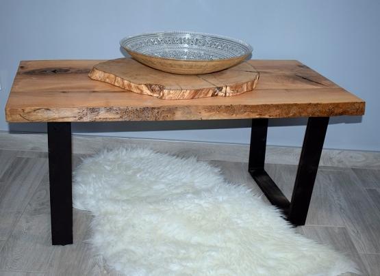 Stolik kawowy z blatem z surowego drewna o nieregularnych krawędziach, wsparty na czarnych, metalowych nogach w kształcie litery V, na blacie leży drewniany plaster i dekoracyjna misa.