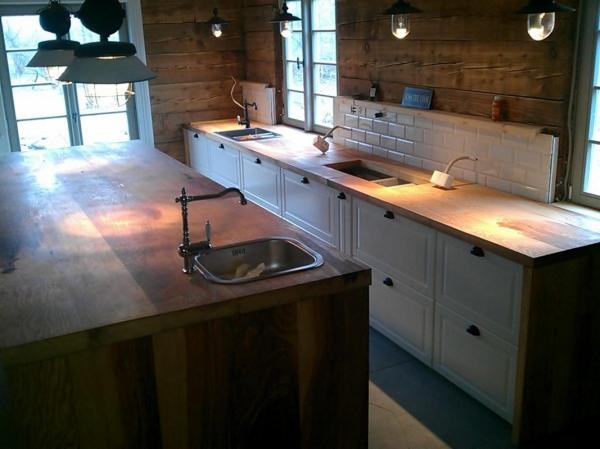 Rustykalna kuchnia z drewnianymi blatami i białymi szafkami, widoczne dwie strefy zlewozmywakowe z armaturą w stylu retro, ściany wykończone drewnianymi panelami, oświetlenie punktowe.