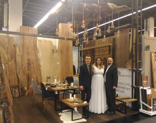 Ekspozycja na targach mebli: stół z blatem drewnianym i krzesłami, tło z dużych desek drewnianych, oświetlenie z żarówek Edisona, oraz trzy osoby w eleganckich strojach pozujące do zdjęcia.