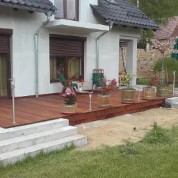 Drewniany taras przy domu z białymi ścianami, kwiaty w donicach, widoczne rolety zewnętrzne w oknach i elementy wykończenia elewacji.