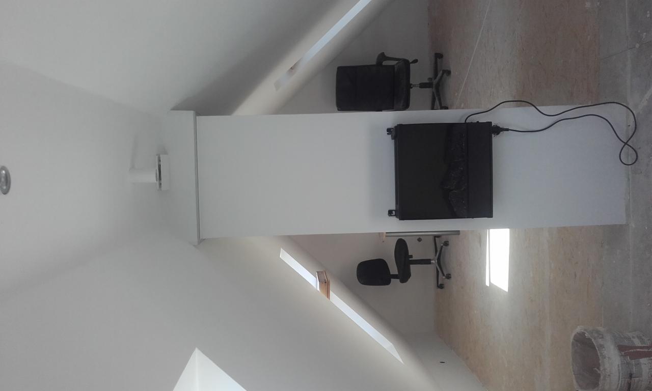 Białe, świeżo pomalowane poddasze ze skosami, z widocznymi oknami dachowymi, dwoma stanowiskami pracy z biurkami i krzesłami, oraz niedomalowaną podłogą i wiadrem po farbie w rogu.