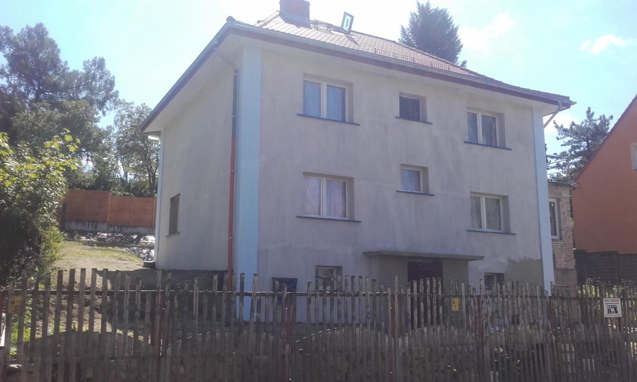 Dwupiętrowy dom z szarą elewacją, częściowo pomalowany na niebiesko przy narożnikach, widoczny przez drewniany płot, z zielenią w tle.