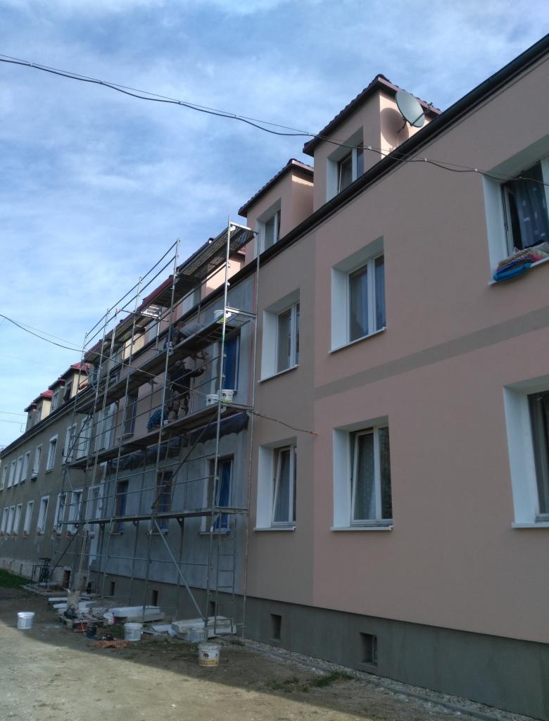 Budynek wielorodzinny w trakcie odnawiania elewacji, widoczne rusztowania, część budynku już pomalowana na beżowo, druga część w stanie surowym, na rusztowaniach pracownicy.