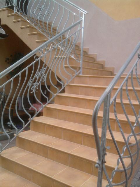 Klatka schodowa wyłożona beżowymi płytkami z metalową, ozdobną balustradą w jasnym odcieniu szarości.