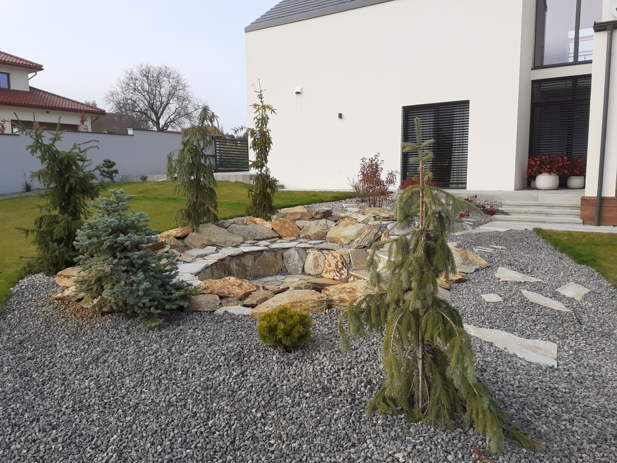 Ogród skalny z małym oczkiem wodnym otoczonym kamieniami, iglaki karłowe i wysypany szarym żwirem na tle nowoczesnego domu z dużymi oknami.