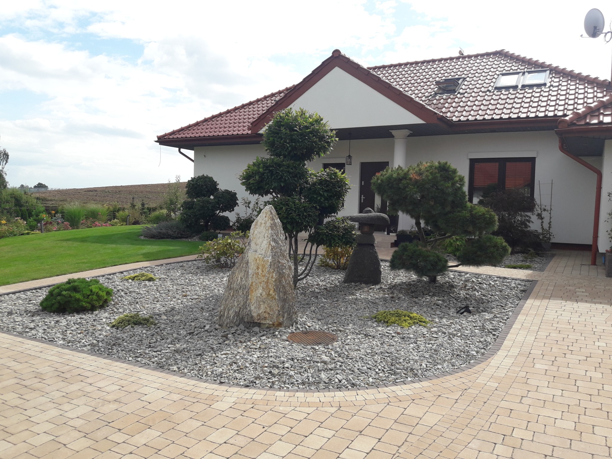 Ogród przydomowy z ozdobnymi krzewami, kamieniami i żwirowym podłożem, widok z brukowanej ścieżki.