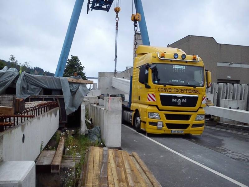 Żółta ciężarówka MAN z napisem 'CONVOI EXCEPTIONNEL' przewozi betonowe elementy konstrukcyjne pod dźwigiem budowlanym.