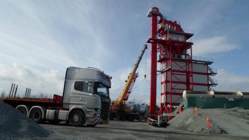 Srebrna ciężarówka Scania z naczepą transportuje elementy czerwonej konstrukcji przemysłowej, widoczny żuraw budowlany, stosy kruszywa na placu budowy, pochmurne niebo.