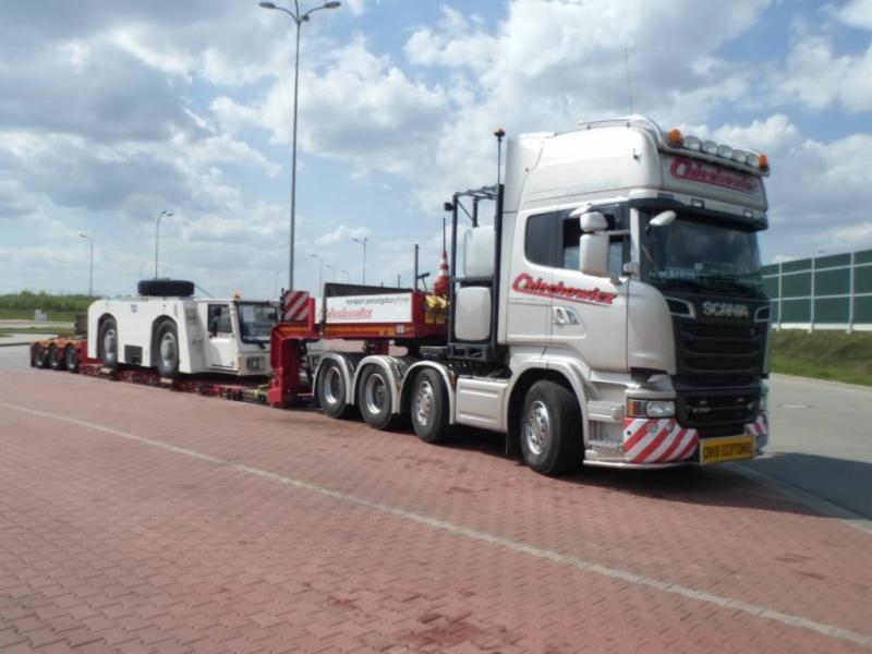 Ciężarówka Scania przewożąca biały pojazd specjalny na niskopodwoziowej platformie, widok z boku na tle pochmurnego nieba.