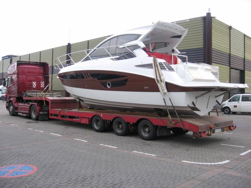 Czerwony ciągnik siodłowy z naczepą niskopodwoziową przewozi luksusowy jacht motorowy w biało-brązowej kolorystyce na tle magazynu.