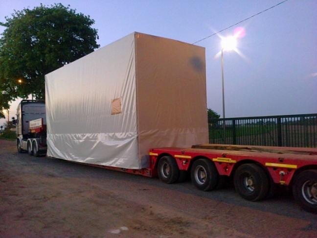 Naczepa z ładunkiem owiniętym białą plandeką, transportowana po drodze gruntowej przy zmierzchu.