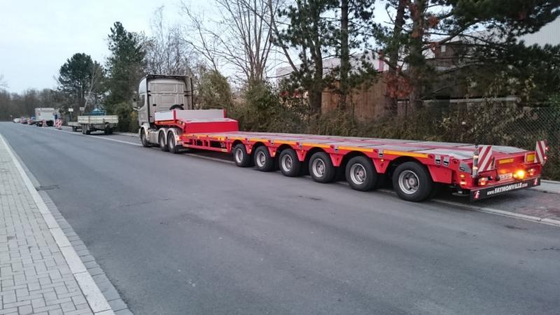 Czerwona naczepa niskopodwoziowa z wieloma osiami, zaparkowana na poboczu drogi, widok z boku, w tle drzewa i budynki.