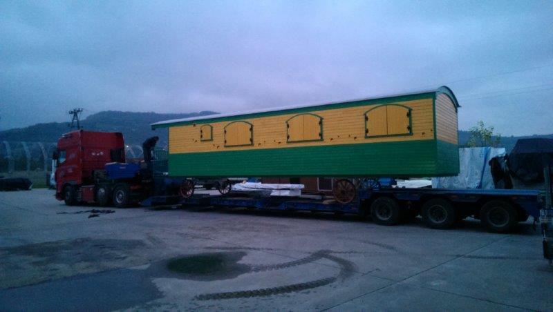 Czerwony ciągnik siodłowy z naczepą przewożący nietypowy ładunek: drewniany, pomalowany na żółto i zielono wóz cygański z zamkniętymi okiennicami, na tle pochmurnego nieba i gór.