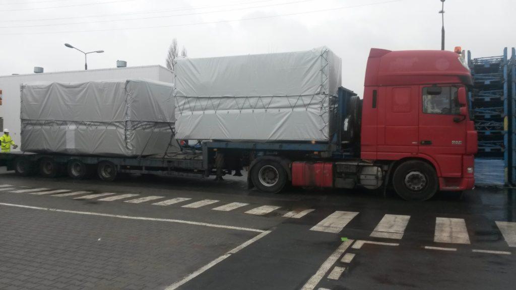 Czerwony ciągnik siodłowy DAF z naczepą przewożącą dwa duże, zabezpieczone plandekami ładunki, widok z boku na tle szarej nawierzchni i budynków przemysłowych.