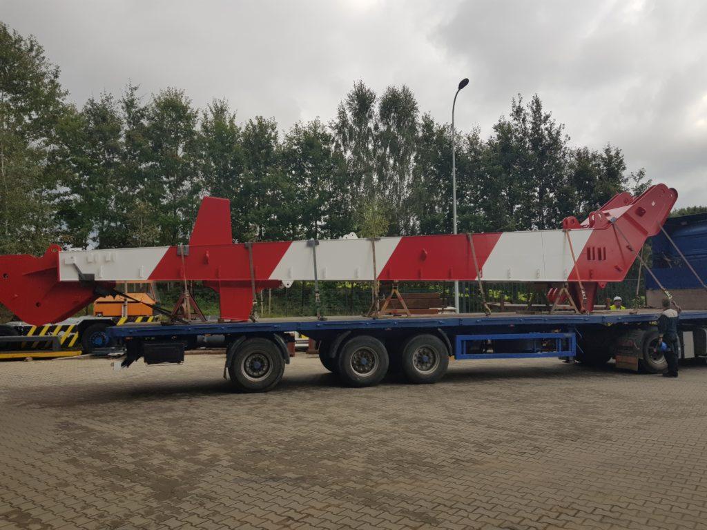 Czerwono-biały element konstrukcyjny dźwigu zabezpieczony pasami na niskopodwoziowej platformie transportowej z trzema osiami, widok z boku na placu manewrowym.