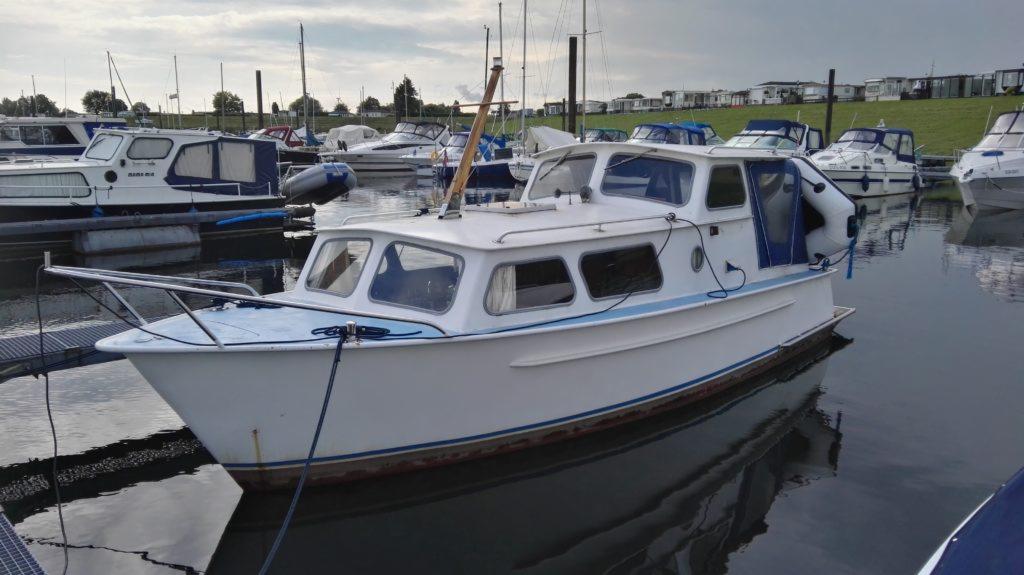 Biała motorówka zacumowana w porcie, widoczne inne łodzie i przyczepy kempingowe na brzegu w tle.