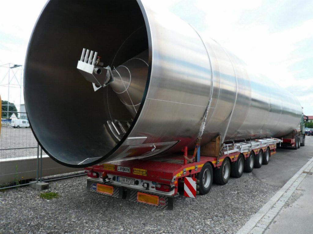 Cysterna ze stali nierdzewnej z mieszadłem wewnętrznym, przewożona na niskopodwoziowej platformie transportowej, widoczna tablica rejestracyjna i elementy mocujące ładunek.