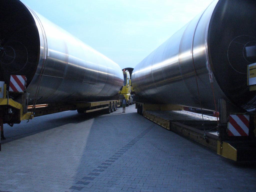 Dwa niskopodwoziowe zestawy transportowe przewożące ponadgabarytowe, metalowe cylindryczne zbiorniki, zabezpieczone pasami, w trakcie postoju na brukowanym placu.