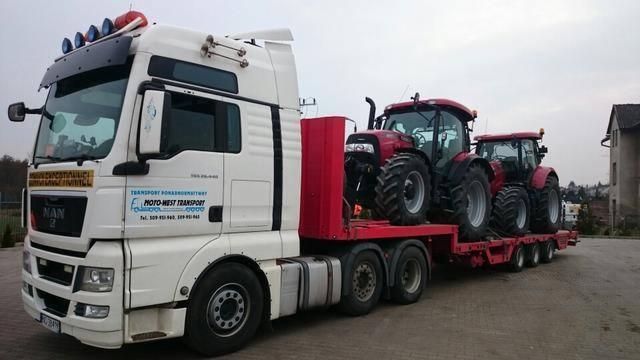 Biała ciężarówka MAN z czerwoną naczepą przewożąca dwa czerwone traktory rolnicze, widoczny budynek w tle.