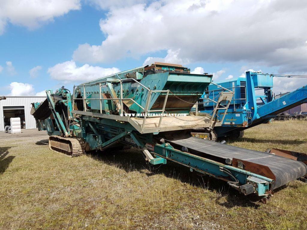 Zielono-niebieska kruszarka mobilna na gąsienicach z taśmociągiem, na trawiastym terenie pod błękitnym niebem z chmurami.