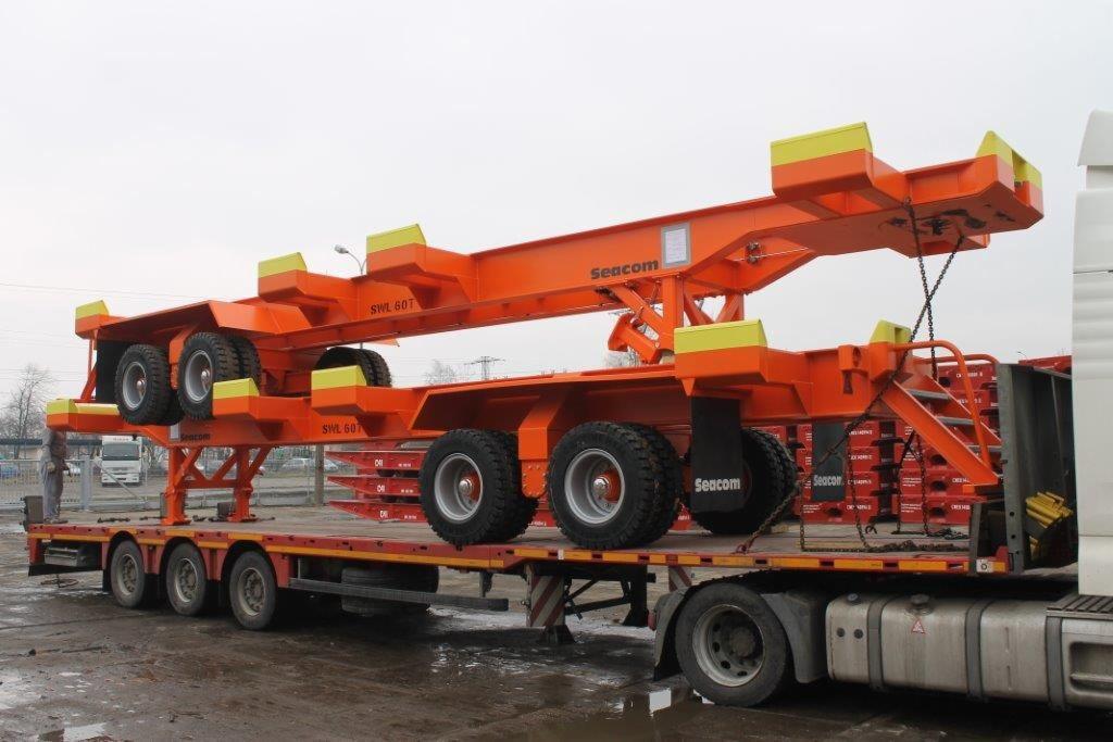Pomarańczowa naczepa niskopodwoziowa z ładunkiem w postaci dwóch modułów transportowych z żółtymi elementami, marki Seacom, na tle szarego nieba i miejskiego krajobrazu.