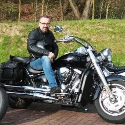 Mężczyzna w skórzanej kurtce i jeansach siedzi na czarnym motocyklu typu trike, trzymając kask; w tle drzewa i trawa.