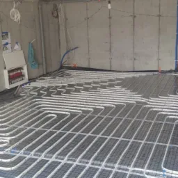 Montaż ogrzewania podłogowego: białe rury układane w pętle na czarnej siatce w surowym pomieszczeniu z betonowymi ścianami, widoczne pionowe rury doprowadzające i rozdzielacz.