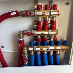 NTS-INSTAL SPÓŁKA Z OGRANICZONĄ ODPOWIEDZIALNOŚCIĄ - Rozdzielacz instalacji wodnej z rurami oznaczonymi do cyrkulacji, umywalki, prysznica, WC i lodówki, zamontowany w białej szafce. Czerwone i niebieskie rury.