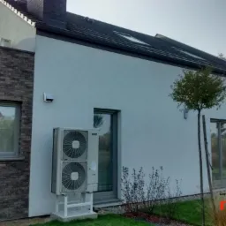 Zewnętrzna jednostka dwuwentylatorowa urządzenia grzewczego zamontowana przy ścianie domu z ceglaną elewacją, widoczna zieleń trawnika i małe krzewy, na ścianie logo firmy.