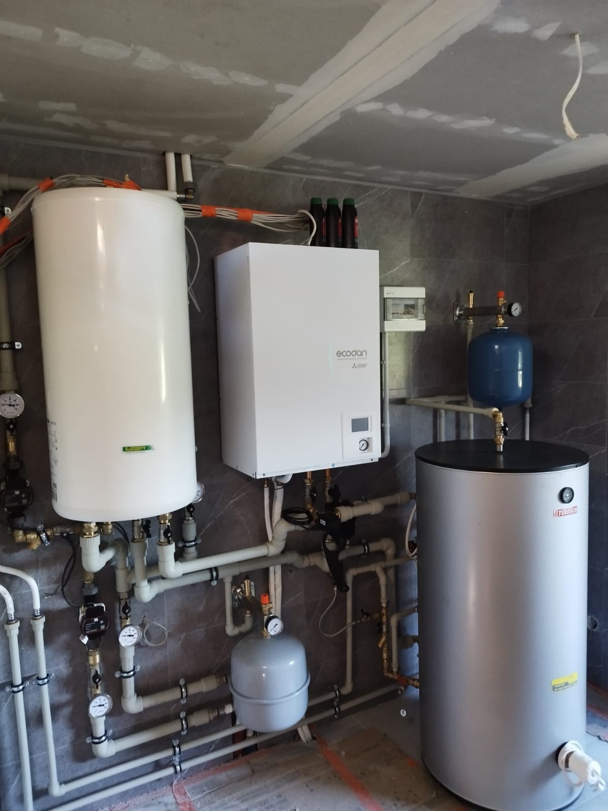 Zestaw urządzeń grzewczych w kotłowni: biały zbiornik buforowy, jednostka zewnętrzna Ecodan Mitsubishi, szary zasobnik ciepłej wody Termica oraz naczynia przeponowe, rury i osprzęt, na tle szarej...