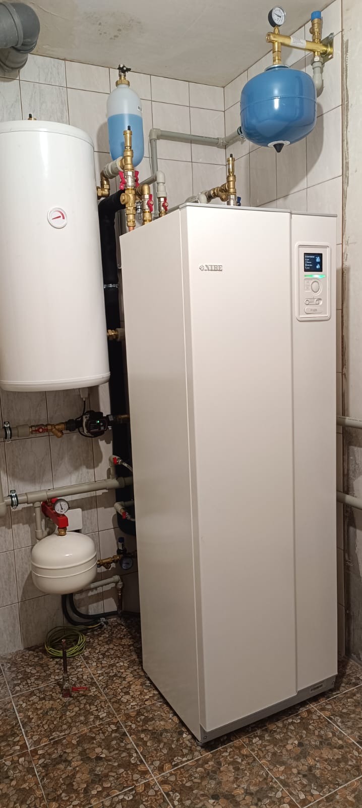 Biała jednostka NIBE z wyświetlaczem, podłączona do instalacji grzewczej z zbiornikiem buforowym i naczyniami przeponowymi, zainstalowana w pomieszczeniu z wykończeniem w płytkach.