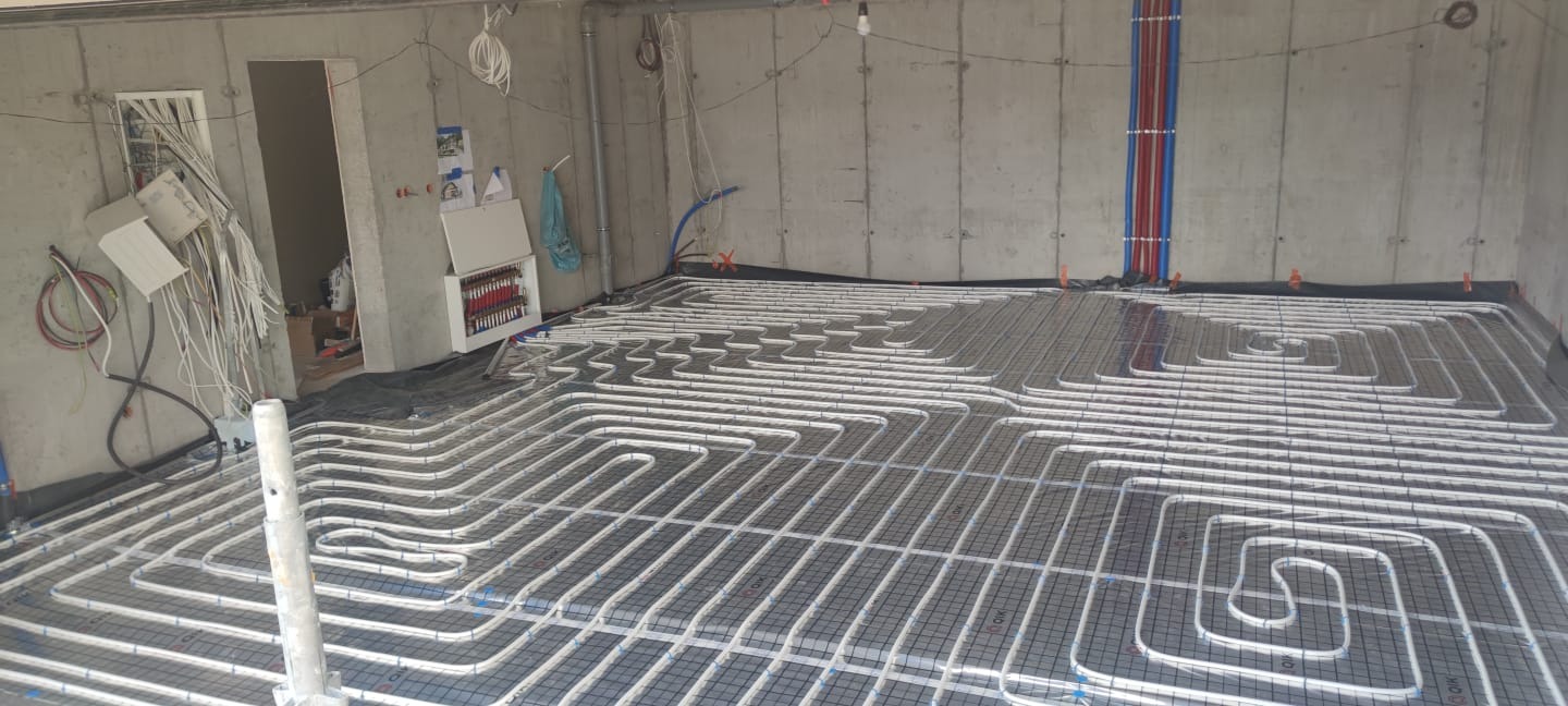 Montaż ogrzewania podłogowego: białe rury układane w pętle na czarnej siatce w surowym pomieszczeniu z betonowymi ścianami, widoczne pionowe rury doprowadzające i rozdzielacz.