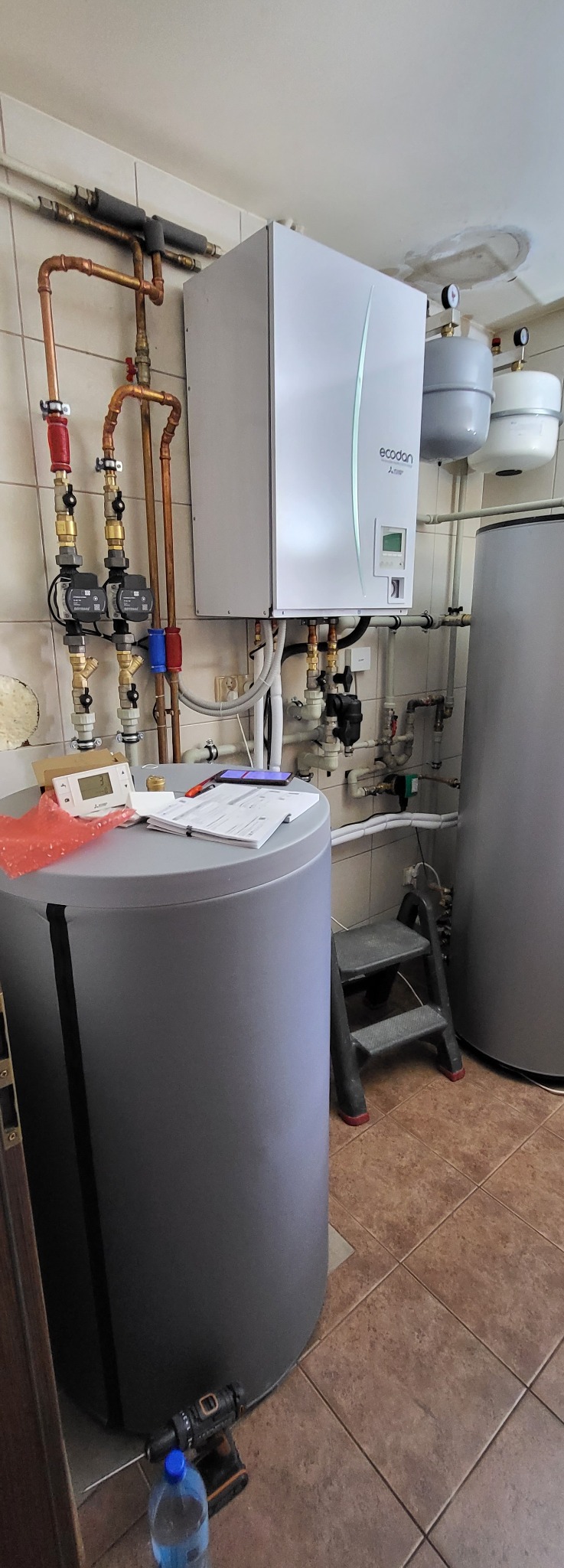 Wnętrze kotłowni z zamontowaną pompą ciepła Ecodan Mitsubishi, zbiornikami buforowymi i rozbudowaną instalacją hydrauliczną z miedzianych rur. Na zbiorniku leżą dokumenty, telefon i sterownik.