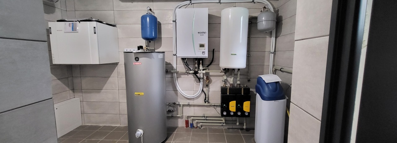 Kompletna instalacja grzewcza w kotłowni: zbiornik, pompa ciepła Ecodan, filtry wody i zasobnik. Rury rozprowadzone wzdłuż ściany z szarych płytek.