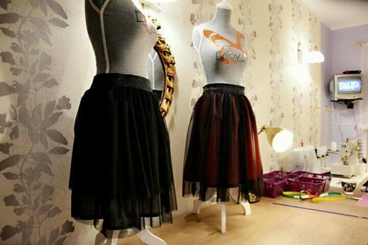 Dwie tiulowe spódnice, czarna i bordowo-czarna, na manekinach w pracowni krawieckiej z tapetą w kwiaty, obok maszyna do szycia i lampka.
