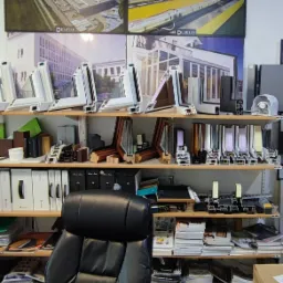 Ekspozycja przekrojów okiennych i drzwiowych różnych materiałów (PCV, drewno, aluminium) na półkach w biurze, z plakatami fabryki okien w tle.